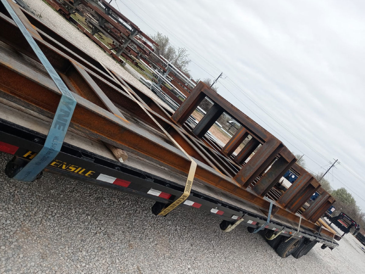Flatbed trailer loaded with rusted steel I-beams secured with tensioning straps at a steel yard