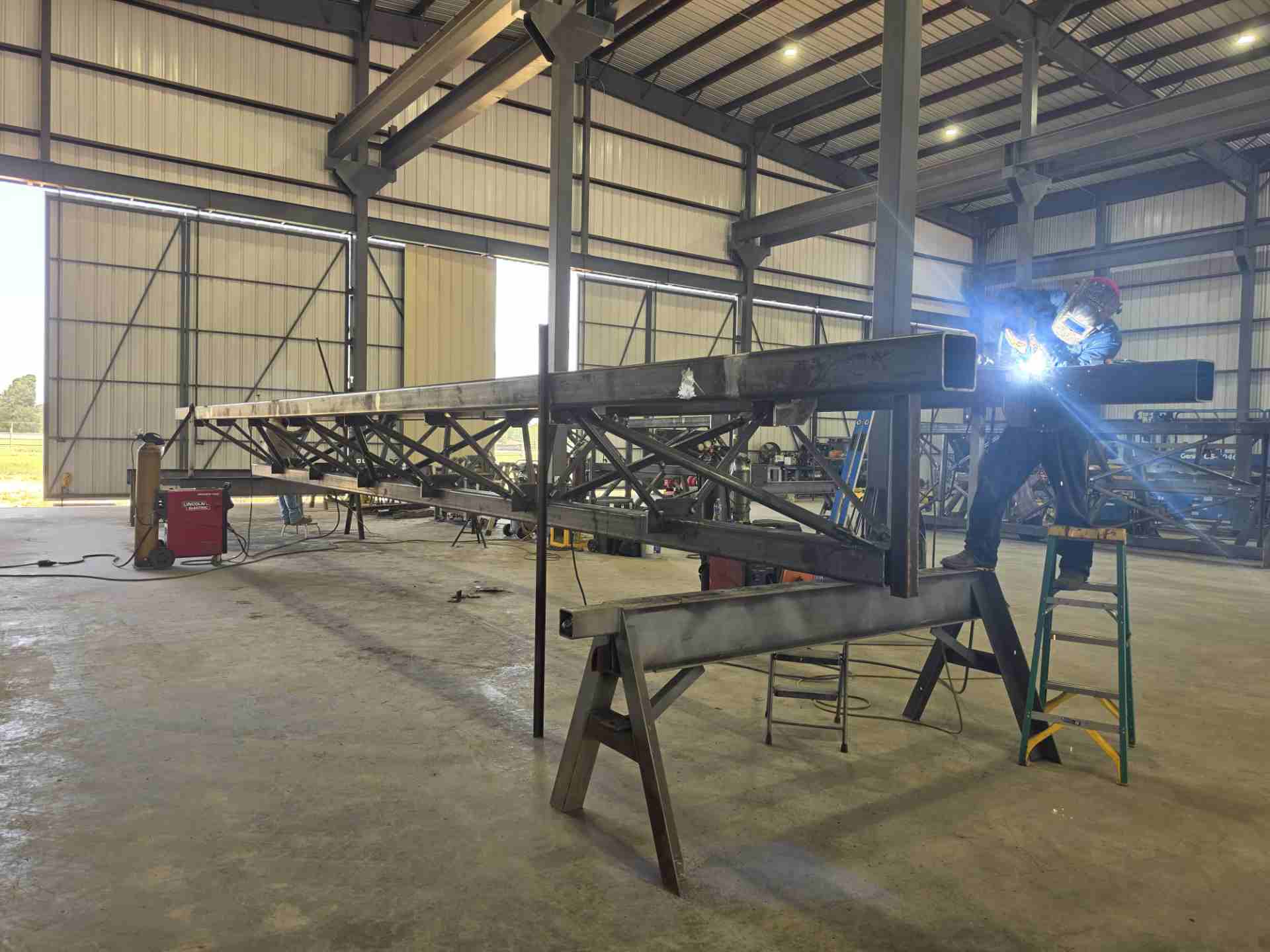 Welder on ladder welding steel truss beam inside industrial metal fabrication shop